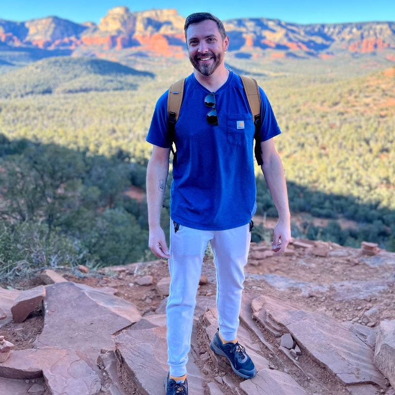 Photo of Logan Bennett on a hike; standing on a rock, smiling, mountains in the distance